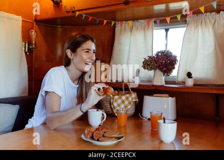 Jolie jeune femme tenant un petit petit pain et riant tout en prenant le petit déjeuner à l'intérieur d'une confortable camionnette Banque D'Images