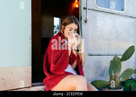 Jolie jeune femme avec une tasse de boisson chaude souriante et vue sur l'extérieur en étant assis sur un campeur en délabeur dans la campagne Banque D'Images