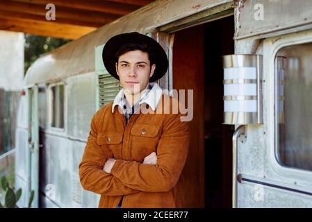 Beau jeune homme aux bras croisés souriant et regardant caméra à proximité de l'entrée d'une caravane âgée Banque D'Images