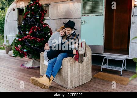 Un jeune homme et une jeune femme joyeux s'embrassant dans un fauteuil près d'un joli arbre de Noël à l'extérieur d'une vieille fourgonnette Banque D'Images