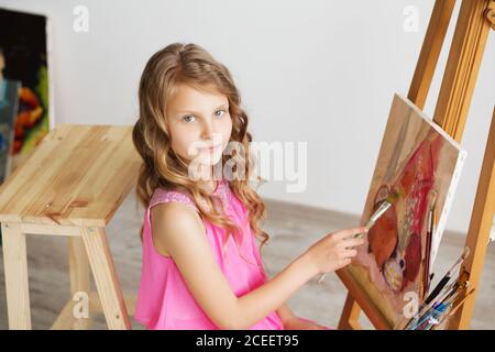 Portrait d'une petite fille qui peint une photo dans un studio ou une école d'art. Le peintre pensif créatif enfant peint une image colorée sur toile avec Banque D'Images