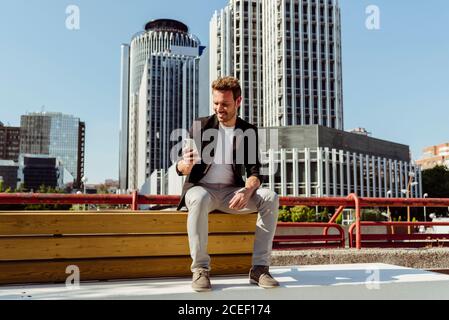 Un beau gars souriant et naviguant sur un smartphone assis sur un banc sur la rue de la ville moderne Banque D'Images