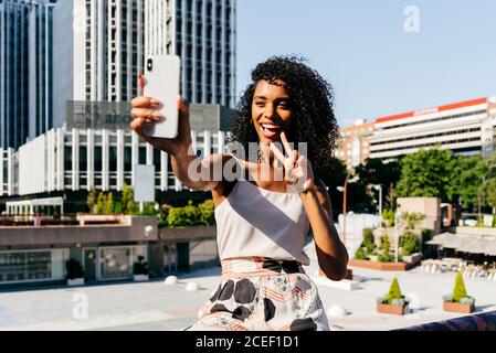 Belle femme afro-américaine en train de rire et de poser pour selfie tout en étant assise sur la frontière sur la rue de la ville le jour ensoleillé Banque D'Images