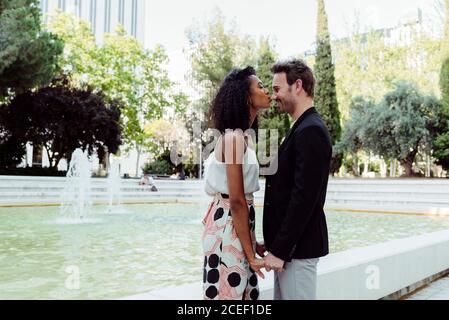 Vue latérale de charmante femme noire et beau homme caucasien s'embrassant tout en se tenant près de la belle fontaine dans le parc Banque D'Images
