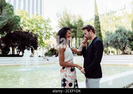 Vue latérale de charmante femme noire et beau homme caucasien s'embrassant tout en se tenant près de la belle fontaine dans le parc Banque D'Images