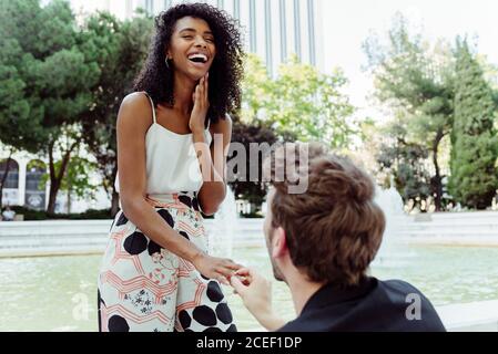 Homme caucasien mettant l'anneau d'engagement sur le doigt de la femme noire tout en lui proposant près de la fontaine dans le parc Banque D'Images