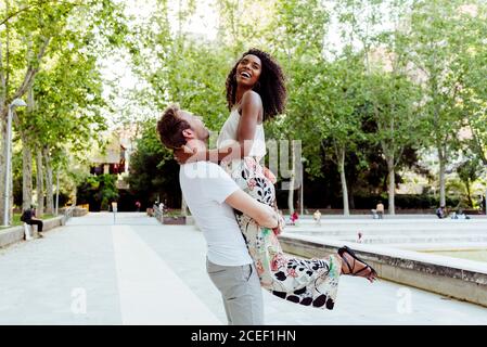 Vue latérale d'un beau caucasien levant une femme afro-américaine en riant sur une allée de parc le jour ensoleillé Banque D'Images
