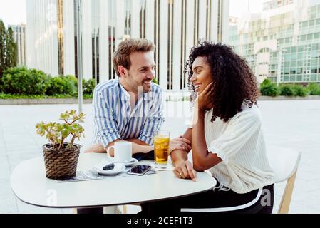 Belle femme afro-américaine et beau homme caucasien souriant et regardant les uns les autres tout en passant du temps dans un café en plein air ensemble Banque D'Images