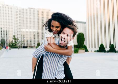 Beau caucasien donnant le pigeyback à la femme afro-américaine tout en s'amusant dans la rue ensemble Banque D'Images