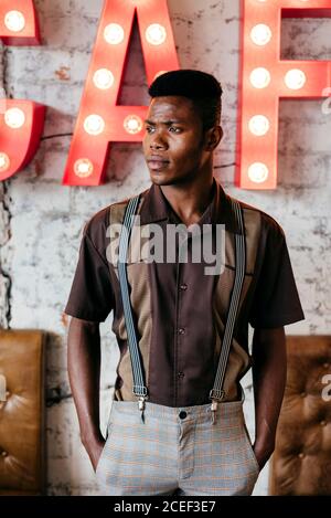 Beau homme africain en pantalon avec bretelles et mise de chemise mains dans les poches debout et regardant l'appareil photo Banque D'Images