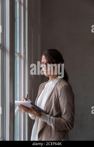 Vue latérale d'une femme adulte dans des lunettes parlant au téléphone et en regardant par la fenêtre pendant que vous vous tenez debout au bureau Banque D'Images