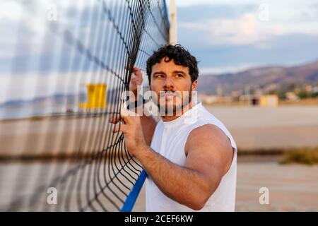 Vue latérale d'un bel homme barbu dans des vêtements de sport touchant le volley-ball net et regarder loin pendant l'entraînement sur la plage Banque D'Images