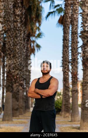 Beau barbu en vêtements de sport en gardant les bras croisés et regardant à la caméra tout en se tenant près des palmiers pendant l'entraînement en extérieur Banque D'Images