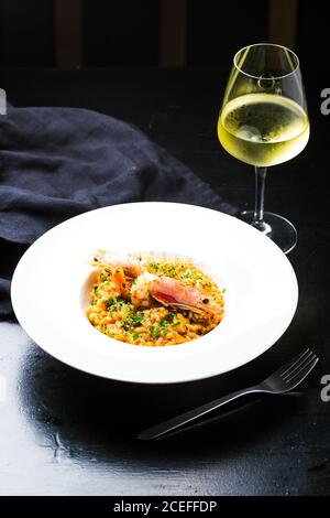 Élégante plaque en céramique blanche avec risotto délicieux aux crevettes placé sur une table en bois sombre avec fourchette et verre de vin blanc Banque D'Images