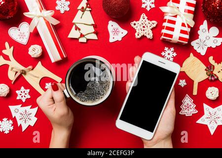 Vue de dessus d'une femme tenant un téléphone dans une main et une tasse de café dans l'autre main sur fond rouge. Décorations de Noël et les jouets. Nouvelle Année holida Banque D'Images