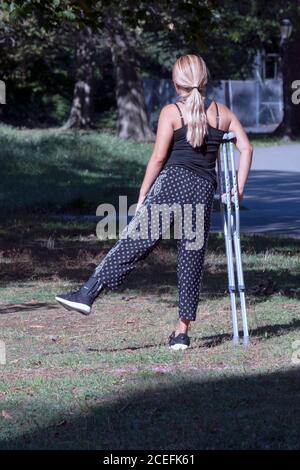 Une jeune femme atteinte d'une cheville étire son autre jambe tout en s'appuyant sur des béquilles. Dans un parc à Queens, New York. Banque D'Images