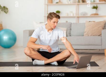 Cours de yoga en ligne. Un homme sérieux travaille dans un ordinateur portable assis sur un tapis Banque D'Images