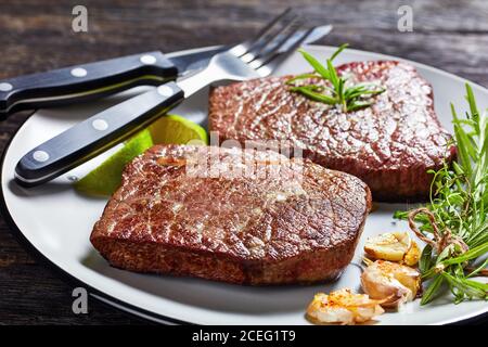 steaks de bœuf frits au romarin, à l'ail et à la chaux sur une assiette avec fourchette et couteau sur une table en bois sombre, vue horizontale depuis le dessus, gros plan Banque D'Images