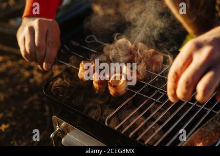 homme préparant du bacon et des saucisses sur des brochettes au feu charbon de bois dans la grille mobile à l'extérieur Banque D'Images