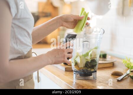 Rognez la vue latérale de la femme en épluchant la moitié de kiwi avec une cuillère et en mélangeant avec d'autres fruits dans le bol mélangeur pour obtenir un smoothie vert Banque D'Images