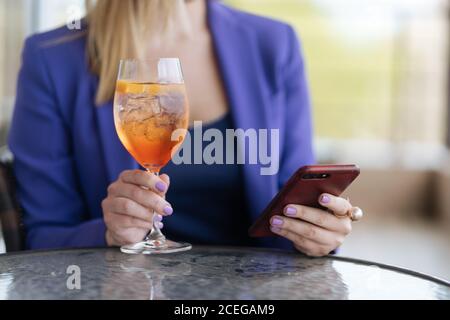 Photo sans visage d'une femme dans une veste violette surfant sur smartphone tout en se relaxant à table avec un verre de délicieux cocktail nordique Banque D'Images