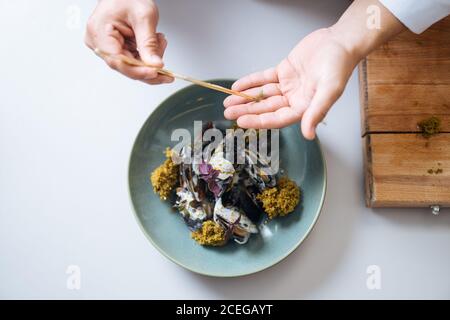 Photo sans visage du chef en uniforme blanc servant des fruits de mer nordiques et verser avec de la sauce dans une casserole debout à la table cuisine Banque D'Images