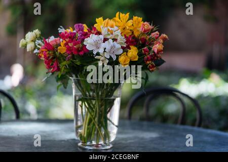 Élégant et coloré fleurs assorties dans un bouquet debout dans un vase en verre avec de l'eau sur une table ronde noire ensoleillée avec des plantes arrière-plan flou Banque D'Images