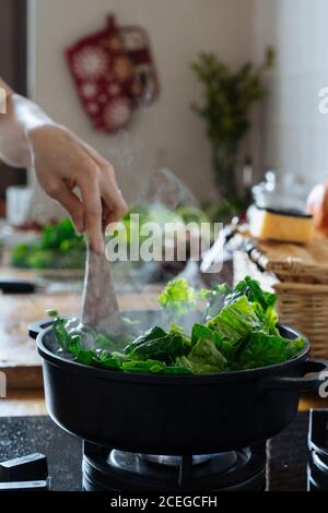 Crop femelle main remuant avec une spatule en bois feuilles de légumes hachées être cuisiné en pot sur cuisinière à gaz Banque D'Images