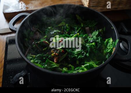 Crop femelle main remuant avec une spatule en bois feuilles de légumes hachées être cuisiné en pot sur cuisinière à gaz Banque D'Images