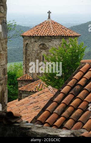 Bâtiment médiéval en pierre avec toit en tuiles rouges et croix chrétienne sur le dessus de l'ensemble architectural ancien à proximité dans le même style avec des arbres surcultivés avec des montagnes et des vallées sur fond Banque D'Images