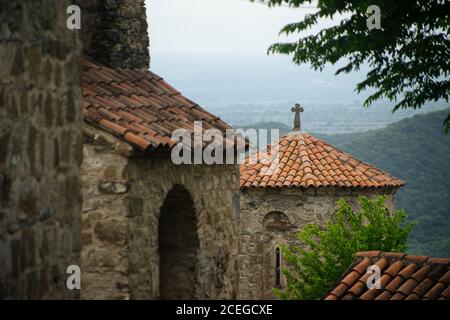 Bâtiment médiéval en pierre avec toit en tuiles rouges et croix chrétienne sur le dessus de l'ensemble architectural ancien à proximité dans le même style avec des arbres surcultivés avec des montagnes et des vallées sur fond Banque D'Images