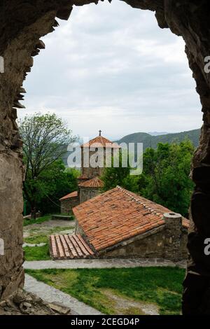 Bâtiment médiéval en pierre avec toit en tuiles rouges et croix chrétienne sur le dessus de l'ensemble architectural ancien à proximité dans le même style avec des arbres surcultivés avec des montagnes et des vallées sur fond Banque D'Images