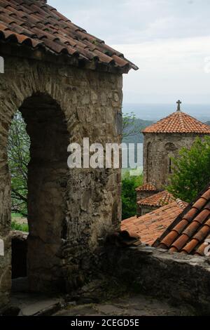 Bâtiment médiéval en pierre avec toit en tuiles rouges et croix chrétienne sur le dessus de l'ensemble architectural ancien à proximité dans le même style avec des arbres surcultivés avec des montagnes et des vallées sur fond Banque D'Images