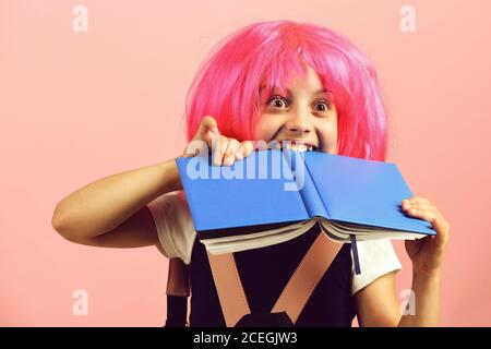 Fille d'école avec la vue surprise isolée sur fond rose. Pupille en uniforme scolaire avec perruque rose. Fille morsures grand livre bleu. Retour à l'école et concept d'éducation. Banque D'Images