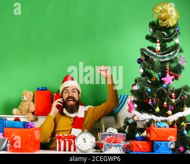 Un homme avec une barbe et un visage joyeux envoie des salutations. Le Père Noël parle sur téléphone portable près d'un sapin sur fond vert. Le Père Noël tient le téléphone portable et met la main vers le haut. Gadgets et concept de célébration Banque D'Images