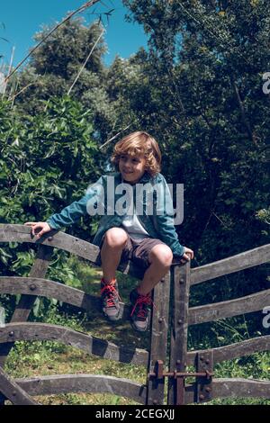 Mignon petit garçon avec des cheveux blond bouclés assis sur petit pont en bois donnant sur la campagne Banque D'Images