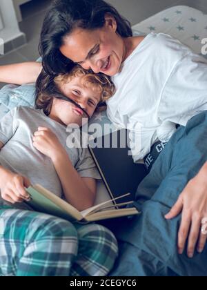 Mignon garçon ayant le plaisir tout en étant allongé sur le lit et la lecture d'un livre intéressant avec l'aide de la mère souriante. Banque D'Images