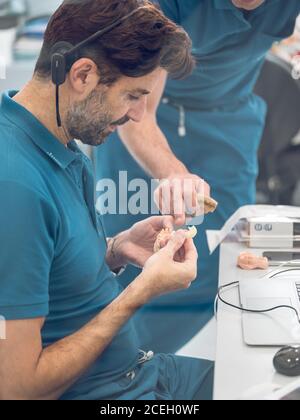 Vue latérale de l'homme beau en uniforme tenant la prothèse et écouter les explications des collègues pendant leur travail en laboratoire dentaire Banque D'Images