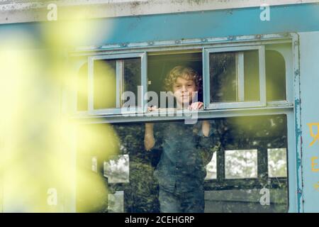 Adorable petit garçon avec des boucles s'amusant à l'intérieur de l'ancien train wagon regardant par la fenêtre et souriant à la caméra Banque D'Images