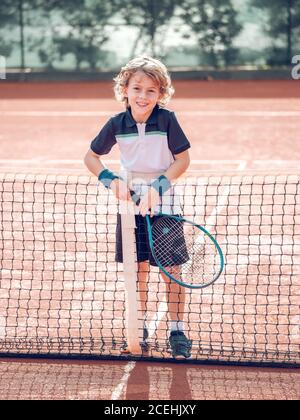 Joyeux petit enfant tenant une raquette de tennis près du filet de tennis court dans la journée ensoleillée Banque D'Images