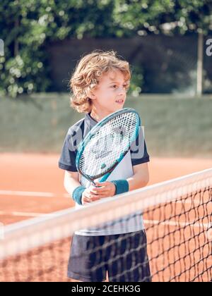 Joyeux petit enfant tenant une raquette de tennis près du filet de tennis court dans la journée ensoleillée Banque D'Images