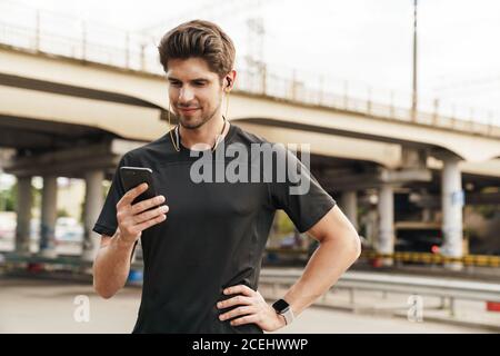 Image d'un jeune sportif heureux dans des écouteurs utilisant le téléphone portable pendant faire de l'activité sur la rue de la ville Banque D'Images