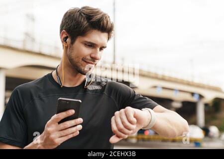 Image d'un sportif heureux avec des écouteurs utilisant un téléphone portable et une montre intelligente pendant votre activité sur la rue de la ville Banque D'Images