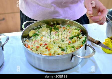 La femme cuit du riz bouilli avec des légumes dans une casserole. Les haricots verts, les poivrons, le maïs, les pois, les oignons et les carottes sont préparés sans perte de vitamines. Banque D'Images