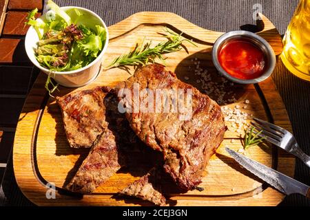 Un délicieux steak de bœuf à la bosse se trouve sur le stand de la planche à découper, accompagné d'une salade fraîche, d'une bière légère en verre, d'une sauce barbecue et d'une branche de romarin. Tableau sur Banque D'Images
