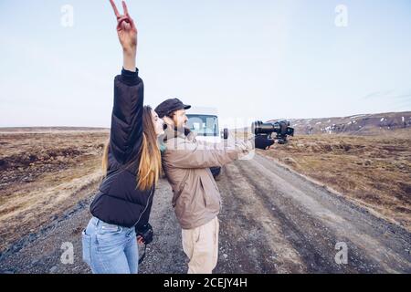 Vue latérale dame souriante avec une main renversant près d'un gars qui prend selfie sur l'appareil photo près de la voiture sur la route entre les terres sauvages En Islande Banque D'Images