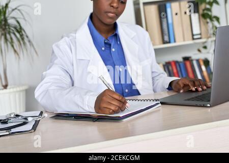 Une femme africaine médecin généraliste porte un manteau blanc en utilisant un ordinateur portable écrit des notes assis au bureau. Banque D'Images