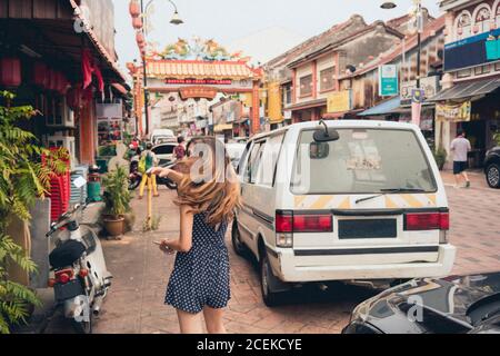 Vue arrière jeune femme mince avec des cheveux venteux en cours Rue asiatique entre bâtiments et automobiles en Malaisie Banque D'Images