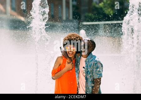 Beau homme afro-américain embrassant la jolie femme et en riant tout en se tenant près de la fontaine ensemble Banque D'Images