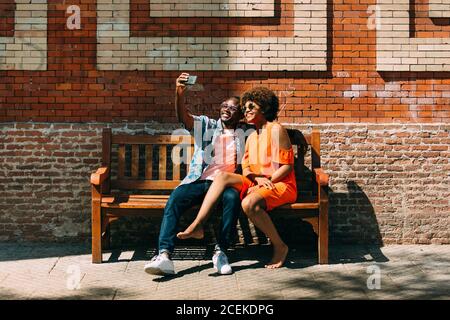 Homme noir et femme pieds nus prenant selfie tout en étant assis sur le banc près du mur de brique Banque D'Images